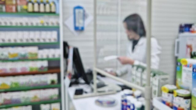 Blurred view inside of pharmacy with female pharmacist at workplace reading medicine prescription. Healthcare and cosmetics industry defocused background