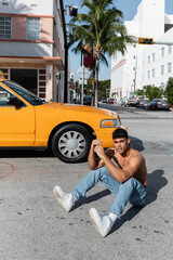 Sexy cuban man with athletic body in baseball cap sitting on road near yellow taxi in Miami