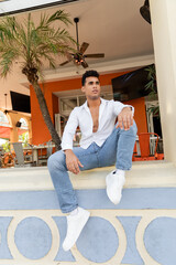 cuban man in white shirt and jeans sitting on parapet of outdoor cafe with palm trees in Miami - obrazy, fototapety, plakaty
