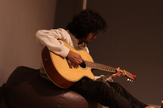 An Indian Boy with long wavy hair playing yellow acoustic guitar while sitting on the bean bag