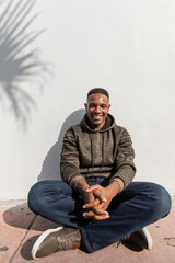 carefree african american man in jeans and hoodie sitting near white wall