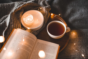 Cup of black tea with burning candle with open paper book on tray in bed over glowing Christmas lights close up. Cozy home atmosphere. Winter holiday season.