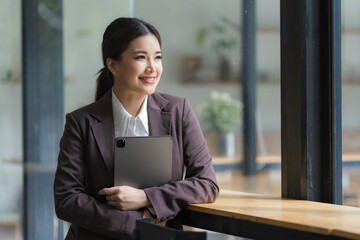 A portrait of a pretty smiley Asian woman standing in a working space holding a tablet and looking away, for business and technology concepts.