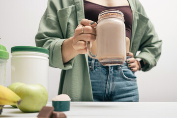 Young woman in jeans and shirt holding glass jar of protein drink cocktail, milkshake or smoothie above white wooden table with measuring spoon of protein powder, chocolate pieces, bananas and apples - obrazy, fototapety, plakaty