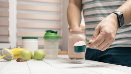 Man holding measuring spoon with protein powder, glass jar of protein drink cocktail, milkshake or smoothie above white wooden table with chocolate pieces, bananas, apples