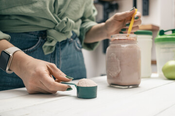 Young woman in jeans and shirt holding measuring spoon with protein powder, glass jar of protein drink cocktail, milkshake or smoothie above white wooden table with chocolate pieces, bananas, apples - Powered by Adobe