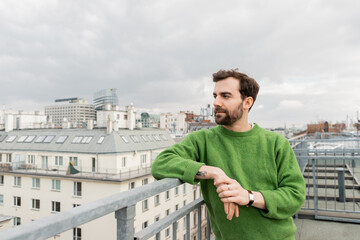 tattooed man in green jumper looking away while standing on rooftop terrace in Vienna, Austria