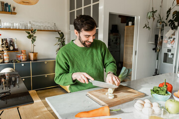 Joyful bearded man in green jumper cutting fresh leek on board at home