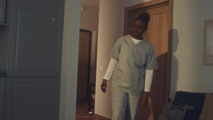 Medium shot of young African American health worker in grey uniform coming home from night shift to family, hugging son browsing on smartphone in kitchen, and kissing husband