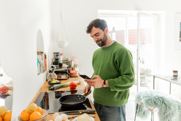 Bearded man smartphone and holding frying pan near eggs, fresh food and butter on worktop