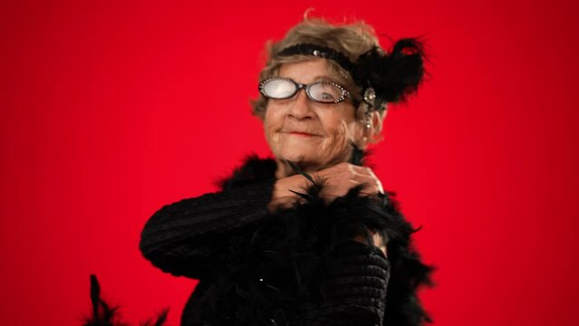 Fun smiling portrait of elderly woman 80s years old walks into frame dancing with boa smiling, isolated on red background studio portrait .