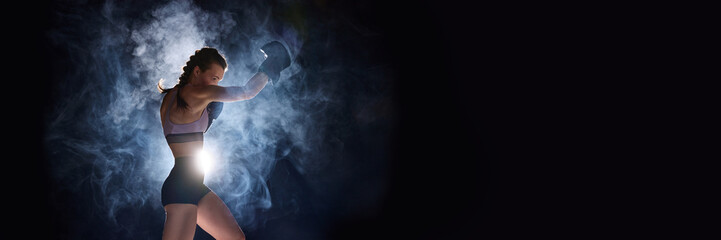 Banner. Flyer. Focused and powerful, female sportsman gearing up for battle, against black studio background in stage smoke. Negative space for text.