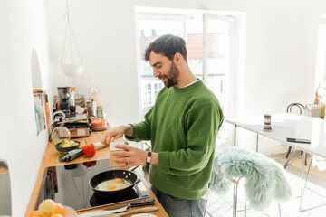 bearded man in green jumper seasoning fried eggs while cooking breakfast in morning