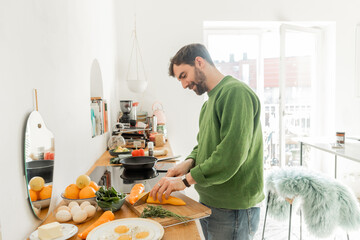 smiling man in green jumper cutting fresh pepper while cooking breakfast in modern kitchen