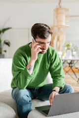 Young brunette and tattooed freelancer in jumper and jeans talking on smartphone and using laptop