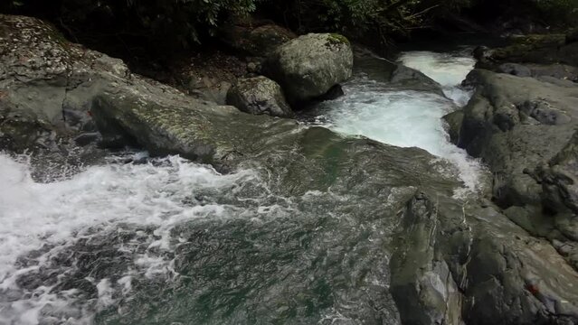 Waterfall cascade falling through stones and rocks in the woodland is filmed from above in slow motion. Tranquil scenery of forest river with strong stream and cliffs at summer. Relaxing view of creek