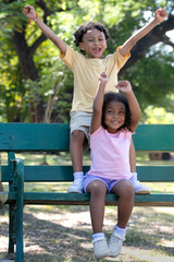 Young sister and her older brother sitting on bench on a bright summer day in the park, have fun by raising your arms, love and bond between brother and sister concept