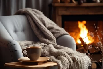 Selbstklebende Fototapeten A mug of hot tea stands on a chair with a woolen blanket in a cozy living room with a fireplace. Cozy winter day © JAYDESIGNZ