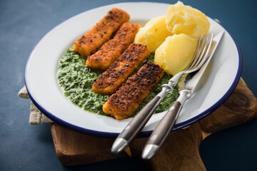 Panierte und gebratene Fischstäbchen mit Rahmspinat und Salzkartoffeln auf Emaille Teller mit Besteck, Holzbrett, Serviette, Spinat Blätter roh und dunkel blau Hintergrund