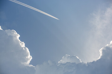 A high flying plane with a trail among epic white clouds and the blue sky.