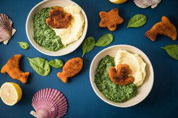 Gebratene Fischstäbchen Figuren als Fisch, Anker, Seestern und Delfin für Kinder mit Rahmspinat und Kartoffel Püree in Schale mit Zitrone, Spinat Blätter roh, Muscheln und dunkel blau Hintergrund