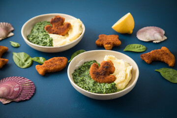 Gebratene Fischstäbchen Figuren als Fisch, Anker, Seestern und Delfin für Kinder mit Rahmspinat und Kartoffel Püree in Schale mit Zitrone, Spinat Blätter roh, Muscheln und dunkel blau Hintergrund
