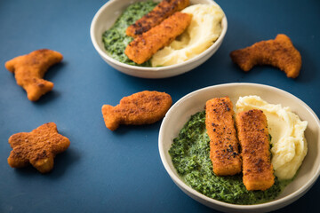 Gebratene Fischstäbchen und Figuren als Fisch, Seestern und Delfin für Kinder mit Rahmspinat und Kartoffel Püree in Schale mit dunkel blau Hintergrund