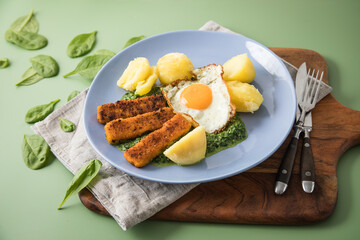 Panierte und gebratene Fischstäbchen mit Rahmspinat, Spiegelei und Salzkartoffeln auf Emaille Teller mit Besteck, Holzbrett, Serviette, Spinat Blätter roh und grün Hintergrund