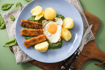Panierte und gebratene Fischstäbchen mit Rahmspinat, Spiegelei und Salzkartoffeln auf Emaille Teller mit Besteck, Holzbrett, Serviette, Spinat Blätter roh und grün Hintergrund