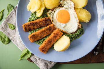 Panierte und gebratene Fischstäbchen mit Rahmspinat, Spiegelei und Salzkartoffeln auf Emaille Teller mit Besteck, Holzbrett, Serviette, Spinat Blätter roh und grün Hintergrund