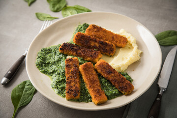 Gebratene Fischstäbchen mit Rahmspinat und Kartoffelpüree auf Teller mit Besteck, Serviette, Spinat Blätter roh und hell grau Stein Beton Hintergrund