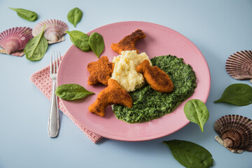 Gebratene Fischstäbchen Figuren als Fisch, Seestern und Delfin für Kinder mit Rahmspinat und Kartoffel Püree auf pink rosa Teller mit Spinat Blätter roh, Muscheln und hell blau Hintergrund
