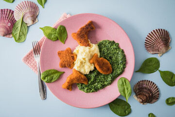 Gebratene Fischstäbchen Figuren als Fisch, Seestern und Delfin für Kinder mit Rahmspinat und Kartoffel Püree auf pink rosa Teller mit Spinat Blätter roh, Muscheln und hell blau Hintergrund © Christin Klose