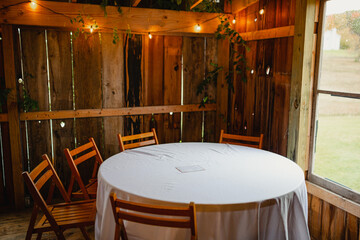 A table covered with a white tablecloth on it with wooden fold out chairs around it in a wedding barn.