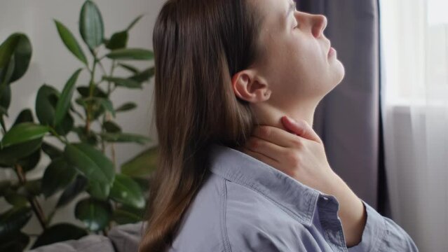 Close up of tired young woman sitting on sofa at home massaging stiff sore neck tensed muscles reduces painful feelings. Shoulder back pain fibromyalgia osteoarthritis senile chronic diseases concept