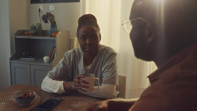 Medium over-shoulder shot of tired black female health worker in medical uniform sitting at kitchen table in morning before shift, drinking coffee with husband, chatting and sharing problem