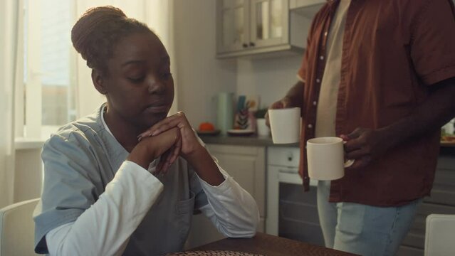 Medium shot of African American health worker in uniform sitting in kitchen with clasped hands, worrying about problems at work, caring husband bringing coffee, hugging, kissing and chatting together