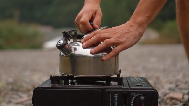 Male hands pouring water from plastic bottle into metal kettle that stands on gas protative stove in nature while traveling.