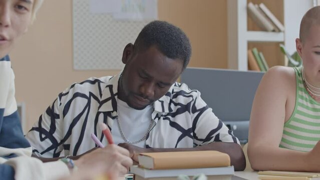 Young Caucasian girl with bald haircut using smartphone while African American guy taking notes and Asian groupmate talking with friend during study day at collegeYoung Caucasian girl with bald haircu