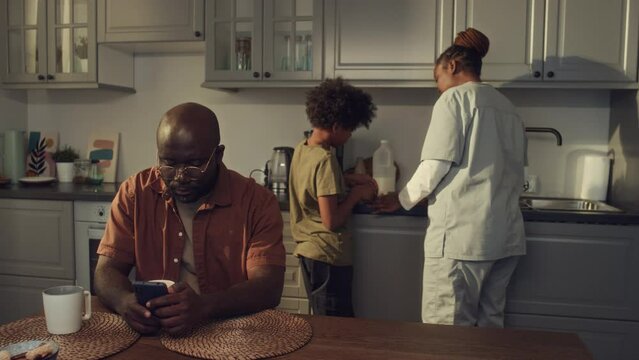 Medium full shot of African American woman in medical uniform standing in kitchen in morning, making sandwiches with son, then carrying them to husband browsing on smartphone and chatting happily