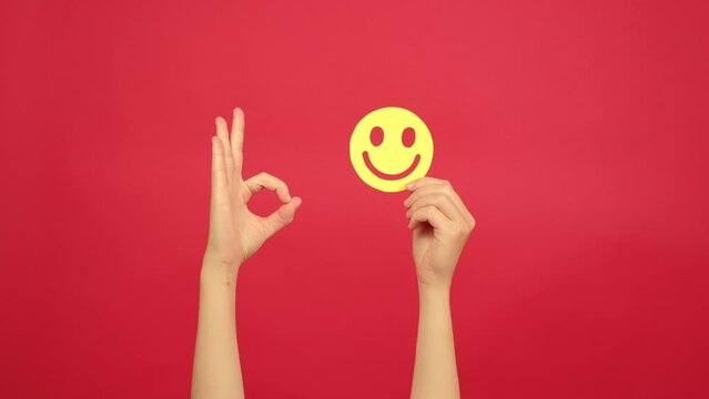 Female hand holding yellow happy smile emoticon, good feedback rating and positive customer review, showing ok sign, satisfaction survey, mental health assessment, isolated over red background wall