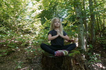 Naklejka premium Little girl in lotus position sitting on the tree stump. Forest meditation concept. Peace, mindfulness and relaxation in nature.