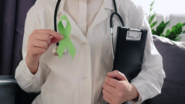 Woman doctor in white coat with stethoscope holding lime green ribbon and clipboard sitting on couch, Mental health awareness, world mental health day, world kidney day, organ donation concept