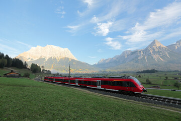 Panoramic view of a train traveling on green fields with Mountain Zugspitze in background on a beautiful sunny day in Lermoos, Tirol, Austria ~ Beautiful summer scenery of idyllic Tyrolean countryside - obrazy, fototapety, plakaty