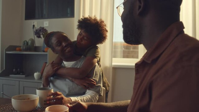 Medium shot of cheerful African American health worker in uniform sitting at table at home, having breakfast, drinking coffee, young son hugging mom, and chatting to husband