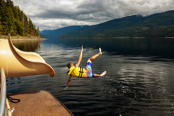 Young boy jumping out of the end of a waterslide on a houseboat on a fall day on Shuswap Lake; British Columbia, Canada