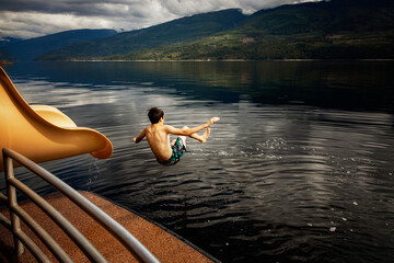 Boy jumping out of the end of a waterslide on a houseboat on a fall day on Shuswap Lake; British Columbia, Canada