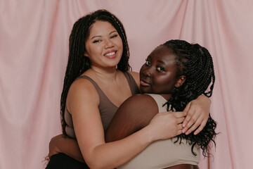 Two beautiful plus size African women in sportswear embracing while standing on fabric background