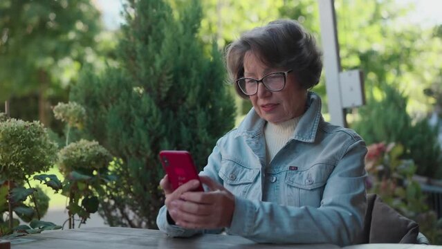 An adult happy gray haired lady using smartphone when sitting on terrace outdoors. Senior woman tapping phone in cafe. Old lady scanned qr code in cafeteria to find meals menu
