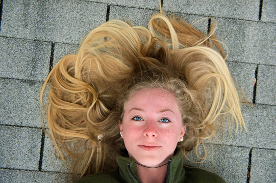 Teenage girl with long blond hair lays on shingled roof; Elkhorn, Nebraska, United States of America